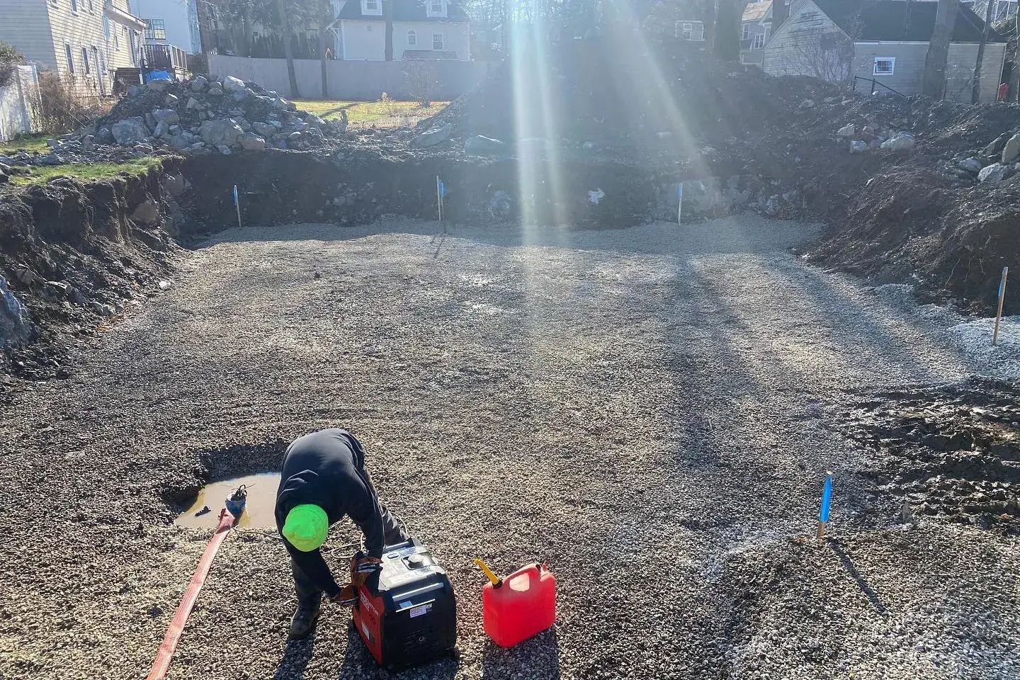 Riverview crew compacting a graded gravel base on a tight residential drainage job, foundation walls and house behind