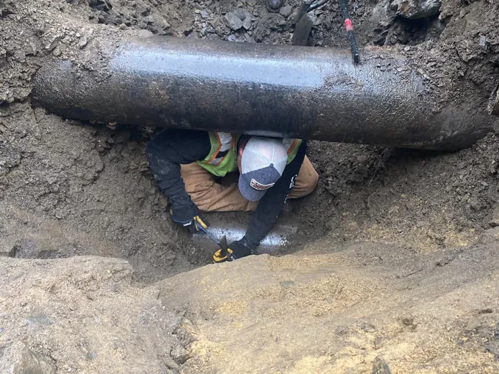 Worker in a hard hat under a large utility pipe in a dig