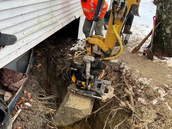 Excavator digging right beside a house foundation with a worker guiding it