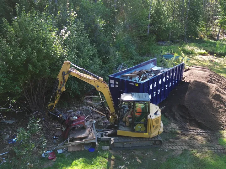 Drone view of an excavator clearing debris into a full dumpster in a backyard