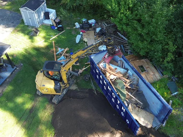 Drone view of an excavator loading debris into a dumpster in a backyard