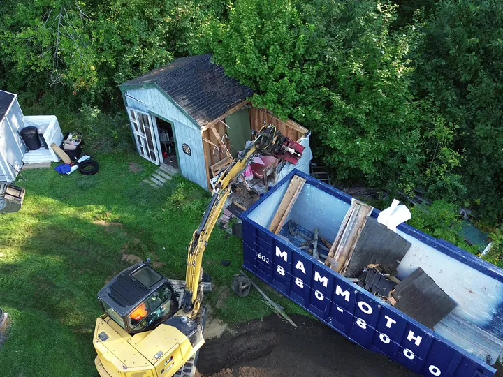 Drone view of an excavator loading debris into a dumpster beside a shed