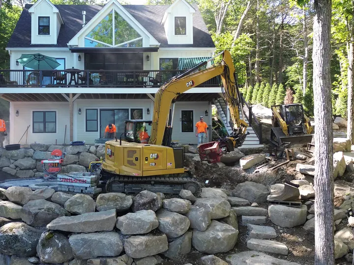 Excavator moving stone in front of a boulder wall below a house
