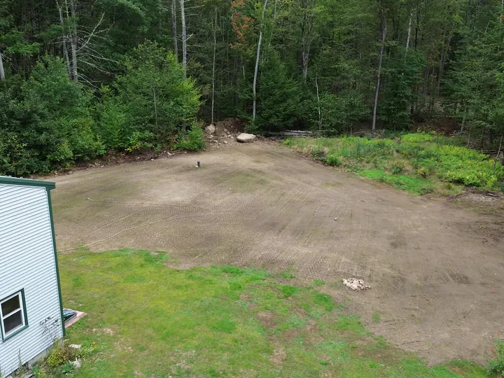 Drone view of a cleared and graded lot beside a house at the edge of the woods