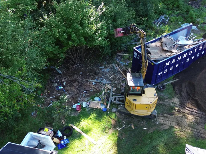 Drone view of a mini excavator beside a loaded dumpster in a backyard