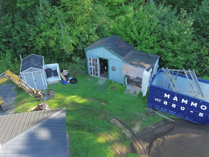 Drone view of a mini excavator tearing down a backyard shed beside a dumpster