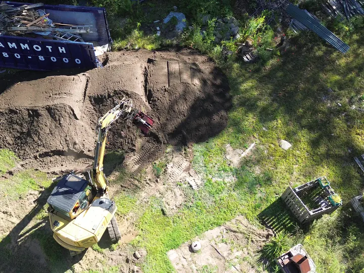 Top down aerial view of an excavator working a pile of fill beside a dumpster