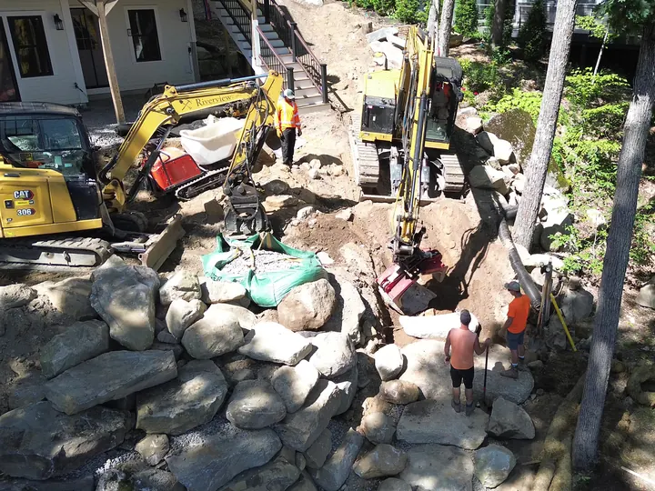 Aerial view of two excavators and crew digging a trench beside a boulder wall