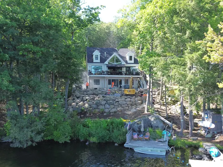 Aerial view of a lakefront house with a boulder wall and an excavator working above the water