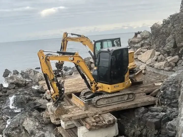 Mini excavator on timber mats over rock beside the ocean