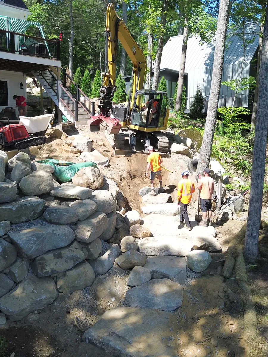 Drone view of a crew building boulder walls and stone steps up a slope below a house, with an excavator at the top