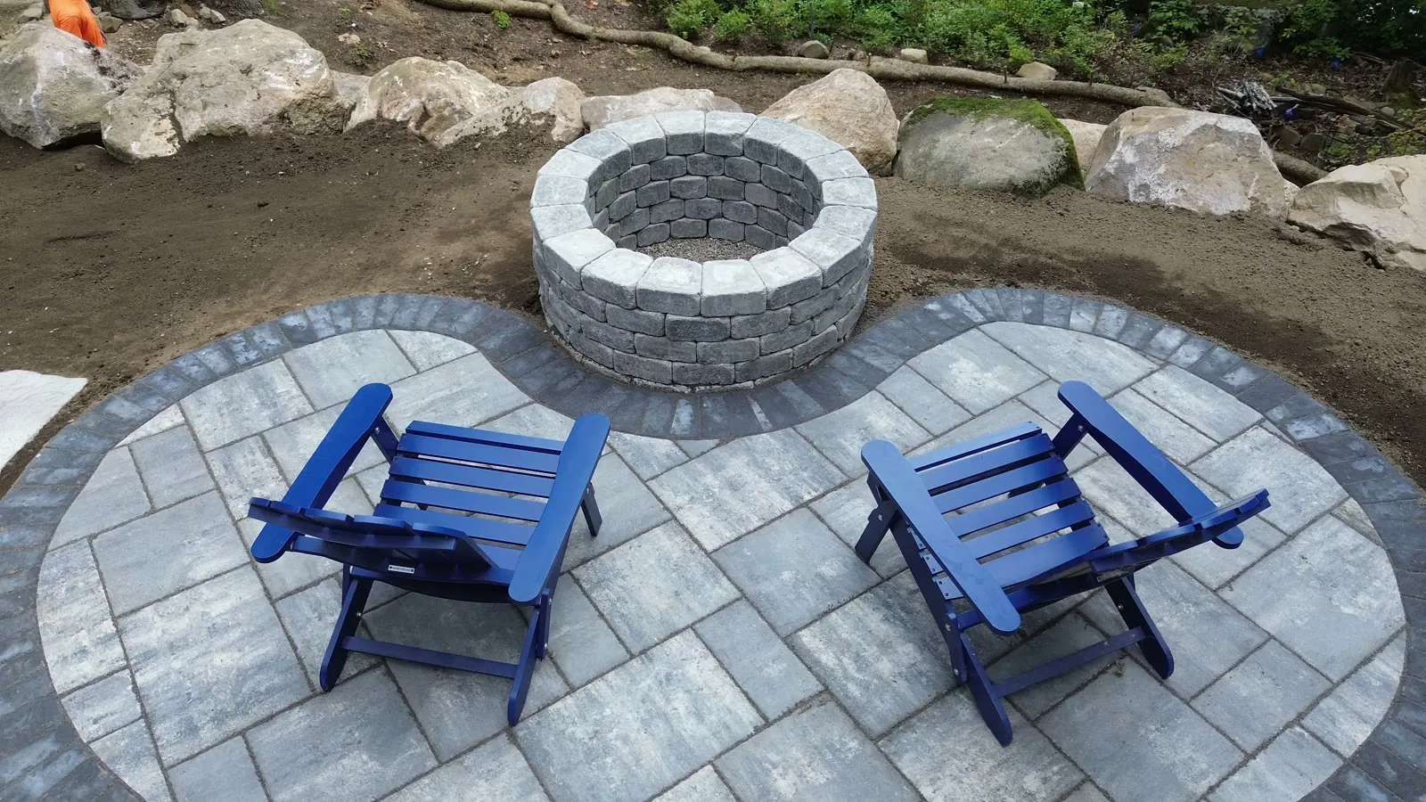 Drone view of a new paver patio with a round block fire pit and two blue chairs, edged with boulders