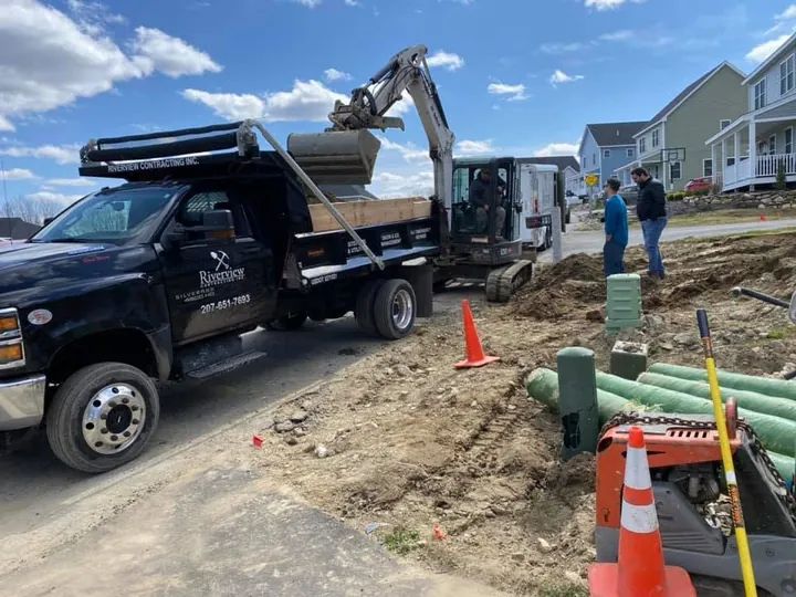 Green pipe stacked beside a trench with a Riverview truck and excavator behind