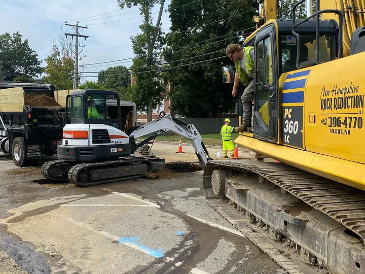 Excavator working a sewer crossing at the edge of a paved road